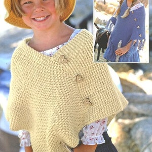 May include: A young person wearing a cream knitted poncho with heart-shaped buttons. The poncho has a V-neck and is knitted in a simple stitch. The person is also wearing a straw hat and a white shirt with floral embroidery.