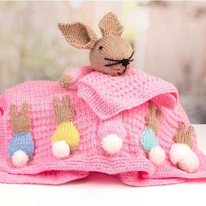 May include: A pink knitted baby blanket with a brown bunny rabbit and four smaller bunny rabbits attached. The blanket has a textured knit pattern and the bunnies have white pom-poms for tails.