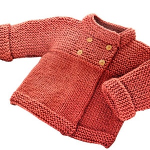 May include: A coral-colored, hand-knitted baby jacket. The jacket features a double-breasted design with three wooden buttons and rolled cuffs. The thick knit creates a textured appearance, perfect for warmth and comfort.