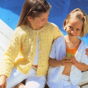 May include: Two young girls wearing knitted cardigans. The girl on the left is wearing a yellow cardigan with white buttons. The girl on the right is wearing a white cardigan with white buttons. Both girls are smiling.