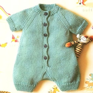 May include: A teal blue knitted baby onesie with a button closure down the front. The onesie has short sleeves and a round neckline.