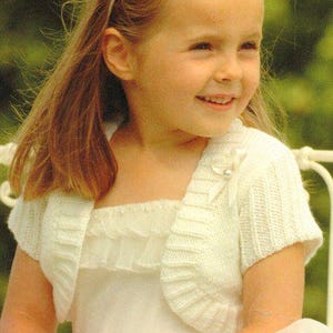 May include: A young girl wearing a white knitted bolero over a white dress with a ruffled neckline. She is wearing a white headband with a flower design.