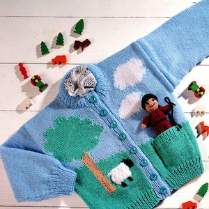 May include: A light blue knitted cardigan with a green, blue and white landscape design. The cardigan features a pocket on the front and a button closure. The design includes a tree, clouds, a sheep and a shepherd figure.