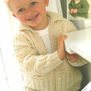 Puede incluir: Primer plano de un niño pequeño con un cárdigan de punto color crema con cremallera. El cárdigan tiene un patrón de punto de cable y se lleva sobre una camiseta blanca. El niño sonríe y se agarra a una mesa. La imagen incluye el texto "Sirdar" y "Incluye tallas para bebé".