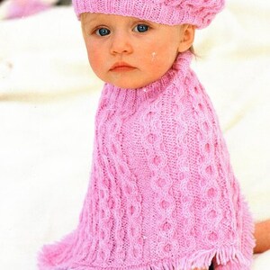 Pode incluir: Um poncho de malha rosa para bebé com um gorro a condizer. O poncho tem um padrão de malha de cabo e uma guarnição de franjas. O gorro tem um padrão de malha simples.