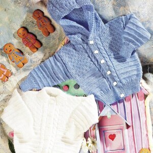 May include: Two knitted baby sweaters, one white and one light blue. The blue sweater has a hood and button closure. The sweaters are laid out on a pink and white striped surface with gingerbread cookies.