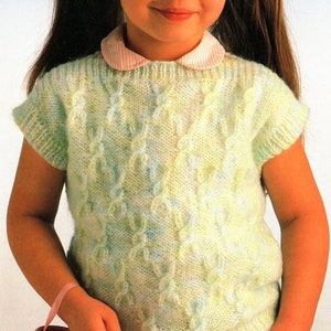 May include: A young girl wearing a light green and blue knitted sweater with a cable knit pattern. The sweater has short sleeves and a round neckline. The girl is wearing a pink and white striped skirt and has her hair pulled back in pigtails.