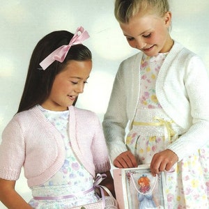 May include: Two girls wearing pink and white knitted cardigans. The girl on the left is holding a pink crocheted purse. The girl on the right is holding a card with a picture of a girl in a white dress.
