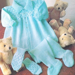 May include: A light blue knitted baby outfit with a button-up front, a collar, and matching booties. The outfit is laid out on a wooden surface with two teddy bears.