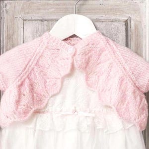 May include: A light pink, knitted bolero sweater with short sleeves, scalloped edges, and a single button closure. The bolero is displayed on a white hanger over a white dress with lace and a bow.