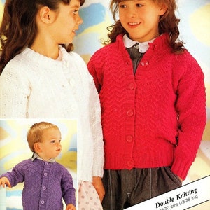 May include: Three children wearing knitted cardigans. The cardigan on the left is white, the cardigan in the center is purple, and the cardigan on the right is pink. The text "Double Knitting 45-70 cms (18-28 ins)" is at the bottom of the image.