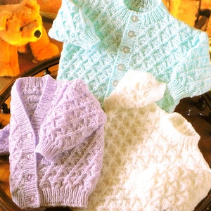 May include: Three knitted baby cardigans in different colors: light blue, lavender, and white. Each cardigan has a diamond pattern and button closure.