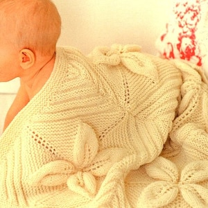 Puede incluir: Una manta de bebé de punto de color crema con un patrón floral. La manta está hecha con un hilo suave y tiene una textura delicada.