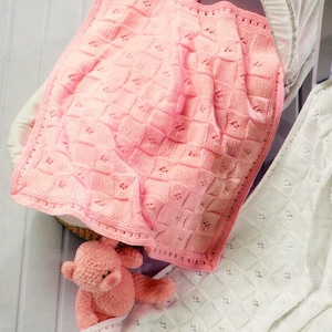 May include: A pink knitted baby blanket with a delicate stitch pattern. The blanket is folded and placed in a white wicker basket. A pink teddy bear is sitting on the blanket.