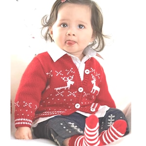 May include: A baby in a red cardigan featuring white reindeer patterns, white buttons, and white edging. The baby is also wearing a white collared shirt, blue jeans, and red and white striped socks. The baby is sticking out their tongue.