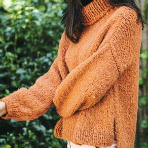 Może przedstawiać: Zbliżenie na kobietę w luźnym, pomarańczowym swetrze z golfem, długimi rękawami i wysokim dekoltem. Sweter wykonany jest z miękkiej, fakturowanej przędzy i ma luźny krój.