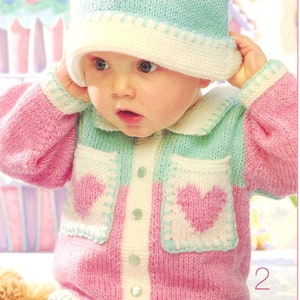 May include: A baby wearing a pink and green knitted cardigan with heart-shaped pockets and a matching knitted hat. The cardigan has a white button placket and a white collar.