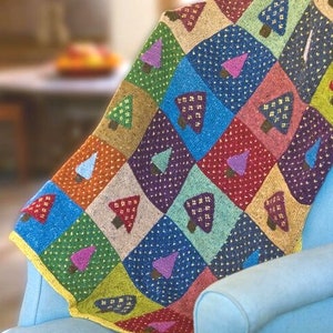 Op de afbeelding: Een kleurrijk gebreid deken met een patchwork ontwerp met verschillende gekleurde vierkanten met kleine bomen in elk vierkant. Het deken is over een blauwe fauteuil gelegd.