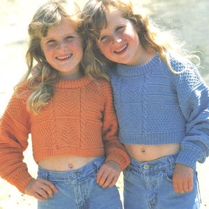 May include: Two girls wearing cropped sweaters. The girl on the left is wearing an orange sweater and the girl on the right is wearing a blue sweater. Both girls are wearing blue jeans.