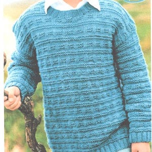 Könnte beinhalten: Ein tealblauer Strickpullover mit strukturiertem Muster. Der Pullover hat einen Rundhalsausschnitt und lange Ärmel. Darunter wird ein weißes Hemd mit Kragen getragen. Der Pullover besteht aus einem weichen, warmen Material.