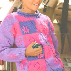 Puede incluir: Una niña lleva un suéter a cuadros morado y rosa con flores bordadas. El suéter tiene un patrón de diamantes en los cuadrados rosas. La niña lleva auriculares y sostiene un pequeño objeto verde.