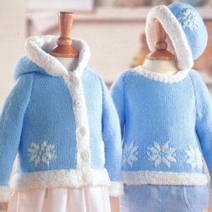 May include: A baby's two-piece outfit in light blue knit with white trim. The set includes a hooded jacket with buttons and a sweater, both featuring snowflake designs. The jacket and sweater have white trim around the edges. The outfit is displayed on wooden mannequins.