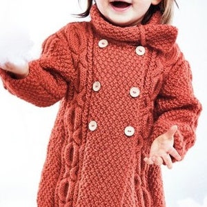 May include: A rust-coloured, double-breasted knitted coat with large, light-coloured buttons. The coat features a textured cable knit design and a high collar. The garment is designed for a child.