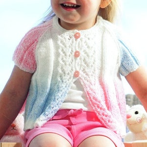 May include: A young girl wearing a white, pink, and blue knitted cardigan with a button closure. The cardigan has a lacy pattern and short sleeves. The girl is wearing pink shorts and is sitting on a wooden surface.