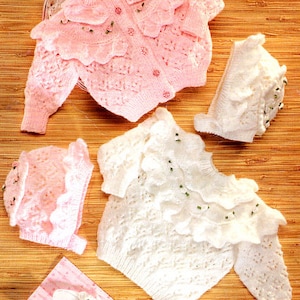 Puede incluir: Dos suéteres de punto para bebé con ribete de volantes y adornos de perlas. Un suéter es rosa y el otro es blanco.