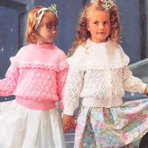 Könnte beinhalten: Zwei Mädchen tragen gestrickte Cardigans mit Spitzenbesatz. Das Mädchen links trägt einen rosa Cardigan und einen weißen Rock. Das Mädchen rechts trägt einen weißen Cardigan und einen Blumenrock.