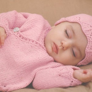 May include: A sleeping baby wearing a light pink knitted outfit, including a jacket with a single button closure and a matching bonnet. The baby is lying on a neutral-colored surface.
