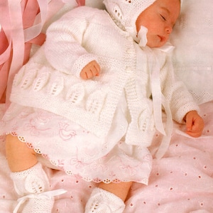 May include: A white knitted baby cardigan with a matching hat, booties, and a white dress underneath. The baby is sleeping on a pink blanket with white polka dots.