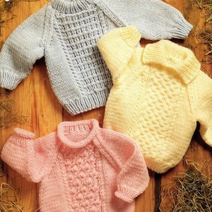 May include: Three knitted baby sweaters in different colors: light blue, yellow, and pink. The sweaters have different stitch patterns and necklines. The sweaters are laid out on a wooden surface with hay.