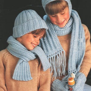 May include: Two children wearing matching light blue knit hats, scarves, and mittens. One child wears a beanie, while the other has a hat that covers the head and ears. The scarves have fringe. The children are wearing tan sweaters.