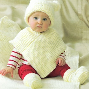 May include: A white knitted baby hat, booties, and poncho set. The hat has ear flaps and the poncho has a simple design.