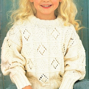 May include: A young girl wearing a white knitted sweater with a diamond pattern. The sweater has a round neckline and long sleeves. The girl is smiling and has blonde hair.