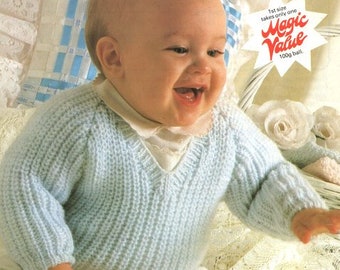 May include: A baby wearing a light blue knitted sweater with a V-neck. The sweater is made with a ribbed stitch. The baby is sitting on a white lace fabric. The text "Patons Fairytale DK" is in the top left corner of the image. The text "1st size takes only one Magic Value 100g ball" is in the top right corner of the image.
