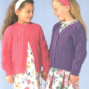 May include: Two girls wearing knitted cardigans. The girl on the left is wearing a pink cardigan with a button closure. The girl on the right is wearing a purple cardigan with a V-neck.