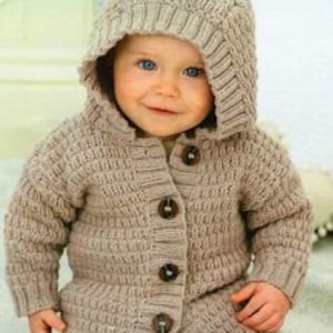 May include: A beige knitted hooded cardigan with bear ears and large wooden buttons. The cardigan is worn over a pair of blue jeans. The baby is smiling.