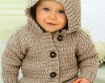 May include: A beige knitted hooded cardigan with bear ears and large wooden buttons. The cardigan is worn over a pair of blue jeans. The baby is smiling.