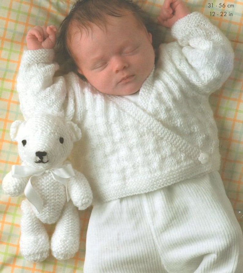 May include: A white knitted baby cardigan with a wrap-around front and a button closure. The cardigan is worn by a sleeping baby, who is holding a white knitted teddy bear. The baby is lying on a checkered blanket.