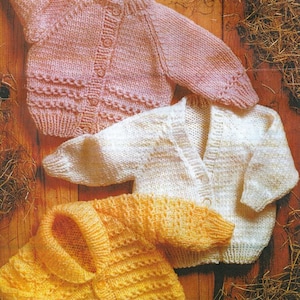 Może przedstawiać: Trzy dzianinowe kardigany dla niemowląt w kolorze różowym, białym i żółtym. Każdy kardigan ma zapięcie na guziki i wzór warkoczowy.