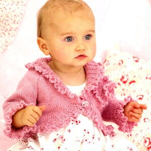 May include: A pink knitted baby cardigan with a ruffled trim around the neckline, sleeves, and hem. The cardigan has a button closure at the front.