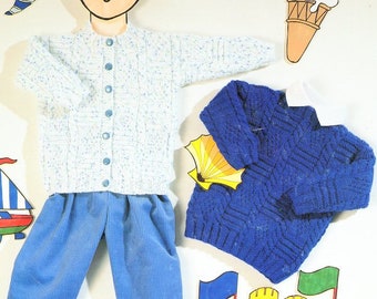 May include: Two knitted sweaters for children. One is white with blue flecks and has buttons down the front. The other is blue with a cable knit pattern. Both sweaters are laid out on a white surface.