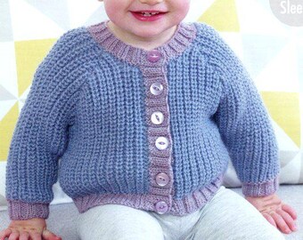 May include: A baby wearing a blue knitted cardigan with a purple trim and buttons. The cardigan is buttoned up and the baby is wearing gray pants. The baby is smiling.