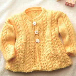 May include: A yellow knitted cardigan with a cable knit pattern and white buttons. The cardigan has a fitted bodice and long sleeves.