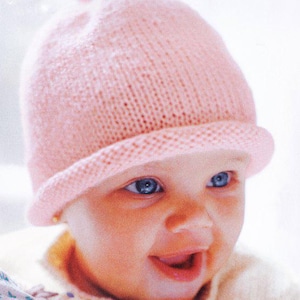 Könnte beinhalten: Ein hellrosa gestrickter Babyhut mit kleinem Rand und Bommel. Der Hut ist aus weichem, strukturiertem Garn gefertigt. Die blauen Augen des Babys sind sichtbar.