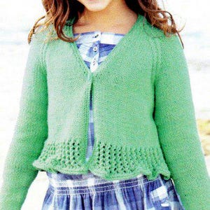 Puede incluir: Un cárdigan de punto verde claro con cuello en V y mangas largas. El cárdigan tiene un dobladillo calado decorativo y se usa sobre un vestido a cuadros azul y blanco. La niña sonríe.