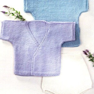 May include: A light blue and lavender knitted baby sweater and white knitted baby shorts. The baby clothes are displayed on a white surface. The baby sweater has a V-neck design. The baby clothes are decorated with sprigs of lavender.