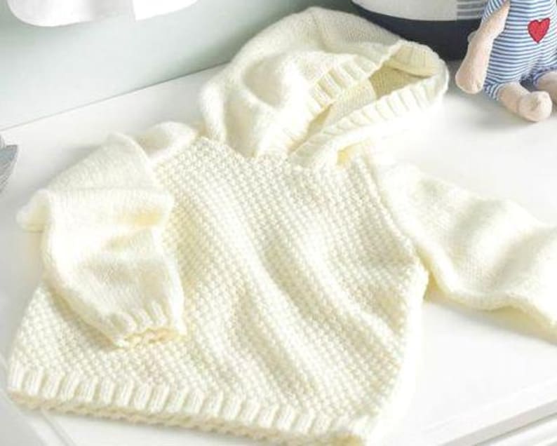 May include: Cream-colored knitted baby hoodie with a textured pattern. The hoodie features a hood, long sleeves, and ribbed cuffs and hem. The soft, warm sweater is designed for a baby or toddler.